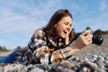 Kayalık bir sahilde neşeli bir kadınla açık hava sahnesi, kamerayla fotoğraf çekiyor, doğanın tadını çıkarıyor, güneş ışığı alıyor, deniz kenarında yaratıcı çekim yapıyor..