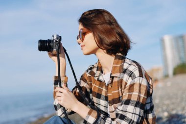 Sahilde sıradan bir fotoğrafçı, elinde kamerayla kompozisyon hazırlıyor. Güneş gözlüğü, ekose gömlek ve mavi gökyüzü deniz kenarında rahat bir portre oluşturur..
