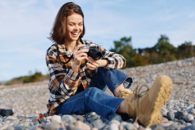 Gündelik kâşif, güneşli bir günde, çakıl taşlarına oturmuş, neşeli ve odaklanmış, rahat bir sahil kenarında seyahat ve doğa fotoğrafçılığını kamerayla yakalar.