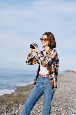 Sahilde kameralı kadın fotoğrafçı, ekose gömlek ve kot giymiş, sahil boyunca ayakta duran, odaklı bir deniz kenarı anı çerçeveleme, görüş açısı, ve parlak bir ışık altında doğal ışık