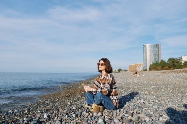 Kayalık plajda bir kadının açık hava portresi, rahat poz, ekose gömlek ve kot pantolon, güneş gözlükleri, sakin denize bakan şehir silueti arkasında, parlak gün ışığı..