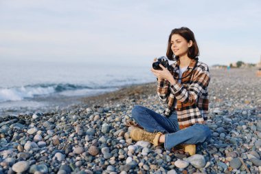 Kayalık bir kıyı şeridinde sıradan bir kadın fotoğrafçı el kamerası, kot ve ekose kıyafet, sakin ruh hali ve okyanus zeminiyle kıyı manzarasını yakalıyor..