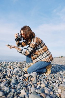 Fotoğrafçı bir çakıl taşı plajında diz çöker, elinde kamera, kayalar ve kıyılar arasında gizli sahneler çeker, doğayı, manzarayı ve kendini adamış uygulamaları taşır..