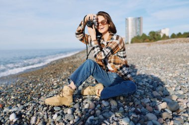 Çakıl taşı plajındaki fotoğrafçı kadın, el kamerasıyla çekim yapıyor, ekose gömlek, kot pantolon ve çizme giyiyor. Güneşli açık hava sahnesi. Deniz meltemi ve uzak binalar.