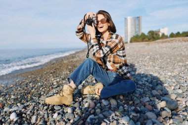 Kayalık bir sahil şeridinde sıradan bir gezgin sahilde fotoğraflar, güneşli hava, kot ve botlar, kameralı gülümseyen kadın, doğayı ve manzarayı keşfederken sahilde hatıraları ve boş zamanları belgeliyor..