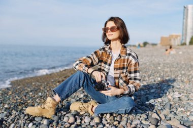 Kayalık bir sahilde el kamerası, rahat bir poz, rüzgarlı saçlar, güneşli bir gün, deniz ufku, kıyı ışığı ve dokuları yakalayan sıradan bir kadın fotoğrafçı..