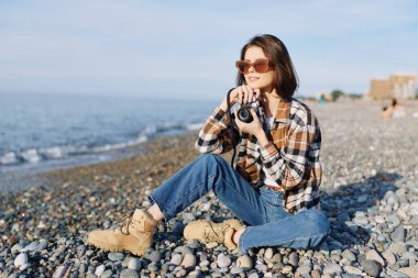 Plaj fotoğrafçısı, açık havada günlük yaşam tarzı deniz taşları güneş gözlüğü, sahil kıyısı, güneş ışığı çakıl taşı plajında ekose kot botlar giyen bir kadınla manzarayı yakalıyor.