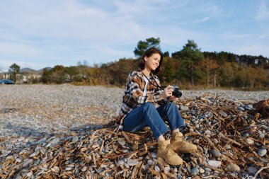 Sahilde kamerası olan bir kadının açık hava portresi, sonbahar manzarası, günlük kıyafetler, yaratıcı fotoğrafçılığa odaklanma, deniz esintisi ve sakin doğa kompozisyonu.
