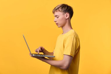 Canlı sarı tişörtlü bir adam dizüstü bilgisayarla etkileşime geçiyor, dijital görevleri yazıyor ve araştırıyor, modern ortamlarda uzaktan çalışmayı, öğrenmeyi ve teknolojiyi gösteriyor..