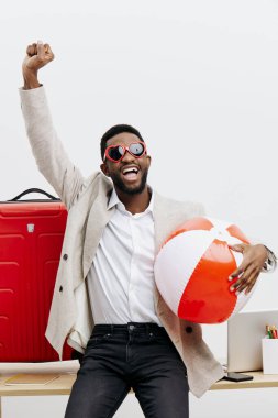 Güneş gözlüklü neşeli bir adam, sıradan giyinmiş, elinde bir plaj topu tutuyor ve modern, parlak bir çalışma alanında duruyor, tatil hazırlığını, boş zamanlarını ve yaz gezilerini ve seyahat heyecanını simgeliyor.