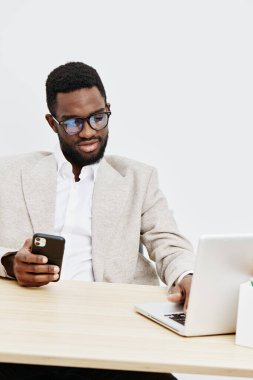 Resmi giyinmiş kendine güvenen siyah bir adam, elinde akıllı bir telefonla masa başında çalışıyor, beyaz arka planda, stüdyo çekimleri yapıyor. Modern çalışma ortamı, teknoloji, iletişim