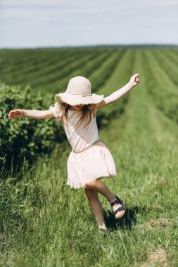 Yazın yeşil bir arazide şapkalı küçük bir kız yürüyor. Arka görünüm