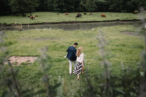 川や森の近くの芝生の上を歩いてハンサムな男とブロンドの女の子 牛や馬の放牧地を背景に手や笑顔を見せる — ストック写真