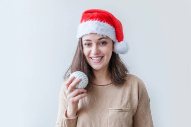 Kırmızı Noel Baba şapkalı güzel bir kız elinde beyaz arka planda izole edilmiş noel ağacı süsü topu tutuyor. Genç kadın portresi, gerçek duygular. Mutlu Noeller ve yeni yıl konsepti