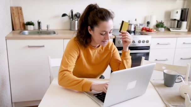 女人在网上购物 用黄金信用卡支付 坐在网上购买笔记本电脑的年轻女孩在厨房室内背景下输入信用卡详细信息 网上购物电子商务概念 — 图库视频影像