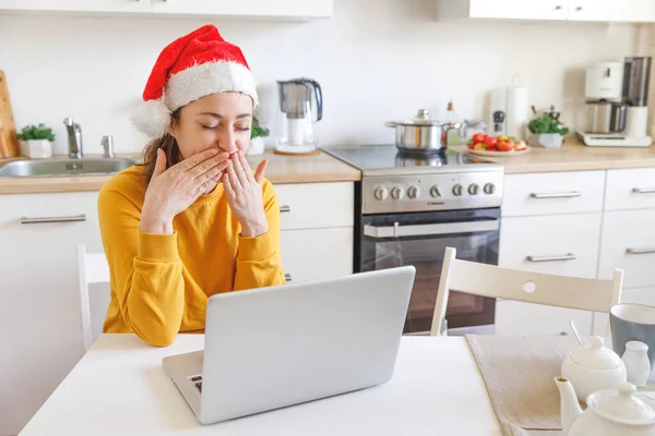 Smiling girl in Santa hat sending air kiss video calling family by webcam. Woman with laptop having virtual meeting chat on holidays sitting on kitchen at home. Happy Christmas and New Year new normal