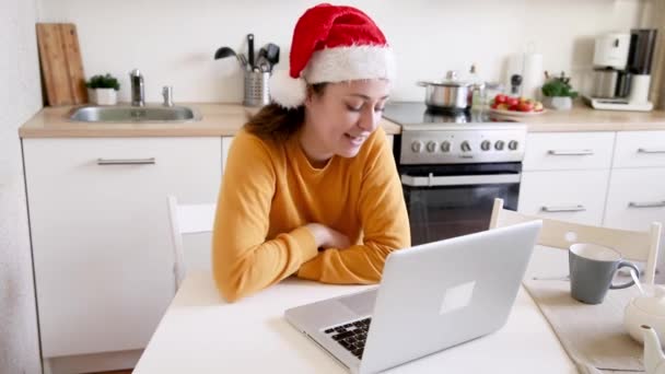 Menina sorrindo vestindo chapéu de Papai Noel acenando mão vídeo chamando a família pela webcam. Mulher com laptop tendo bate-papo reunião virtual em férias sentado na cozinha em casa Feliz Natal e Ano Novo novo normal — Vídeo de Stock