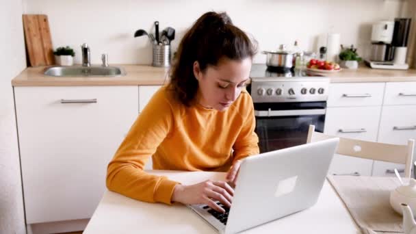 Mobiles Büro zu Hause. Junge Frau, die zu Hause in der Küche sitzt und am Laptop arbeitet. Lifestyle-Mädchen studieren oder arbeiten in Innenräumen. Freiberufliches Quarantäne-Konzept. — Stockvideo