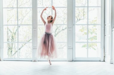 Dans sınıfındaki genç klasik balerin kız. Güzel, zarif balerin, bale pozisyonu pembe etekli, beyaz ışıklı salonun geniş penceresinin yanında.