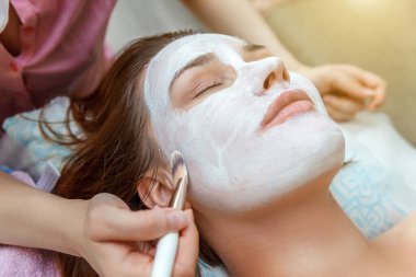 Güzellik salonundaki genç, esmer kadının beyaz kremayla yüz bakımı gördüğü yakın plan. Yüz masajı. Kaplıca ve vücut bakımı. Cilt temizleme kozmetik rahatlama konsepti