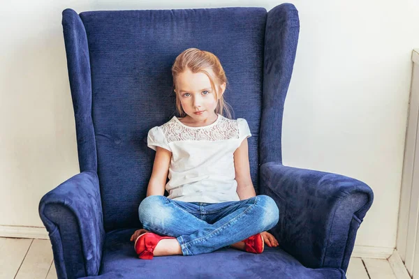 Sweet little girl in jeans and white T-shirt sitting on modern cozy blue chair relaxing in white bright living room at home indoors. Childhood schoolchildren youth relax concept