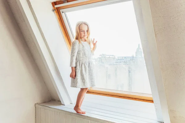 家の中で明るい光のリビングルームで窓の上に立って白いドレスの小さなかわいい甘い笑顔の女の子。子供の頃の若者のリラックスコンセプト. — ストック写真