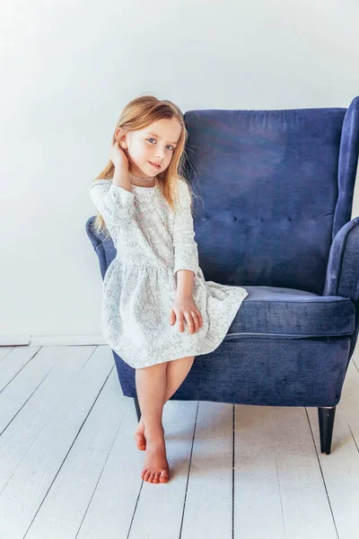 Lief klein meisje in witte jurk zittend op moderne gezellige blauwe stoel ontspannen in witte heldere woonkamer thuis binnen. Jeugd school kinderen jeugd ontspannen concept. — Stockfoto