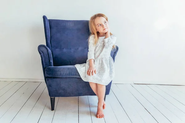 Dulce niña en vestido blanco sentado en la silla azul acogedor moderno relajarse en blanco brillante sala de estar en casa en el interior. Los escolares de la infancia los jóvenes relajan el concepto. — Foto de Stock