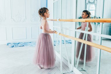 Dans sınıfındaki genç klasik balerin kız. Güzel, zarif balerin, bale pozisyonu pembe etekli, beyaz ışıklı salonda büyük aynanın yanında.