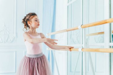 Dans sınıfındaki genç klasik balerin kız. Güzel, zarif balerin, bale pozisyonu pembe etekli, beyaz ışıklı salonda büyük aynanın yanında.