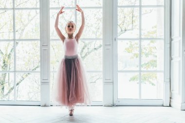 Dans sınıfındaki genç klasik balerin kız. Güzel, zarif balerin, bale pozisyonu pembe etekli, beyaz ışıklı salonun geniş penceresinin yanında.
