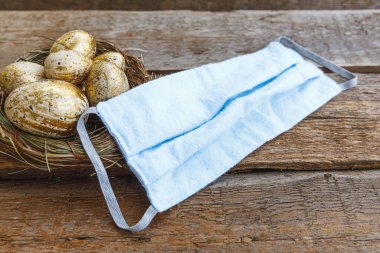 Paskalya karantinası koronavirüs konsepti. Mutlu Paskalyalar, tatil hazırlıkları. Yuvasında altın madalyalı paskalya yumurtaları ve tahta arka planda yüz koruyucu maske. Düz görünüm kopyalama alanı