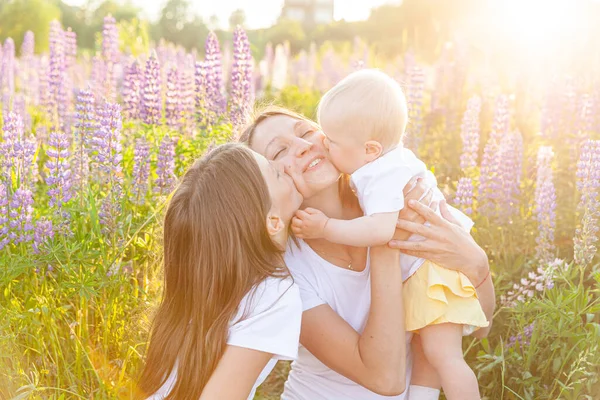 Young mother embracing her kids outdoor. Woman baby child and teenage girl sitting on summer field with blooming wild flowers green background. Happy family mom and daughters playing on meadow