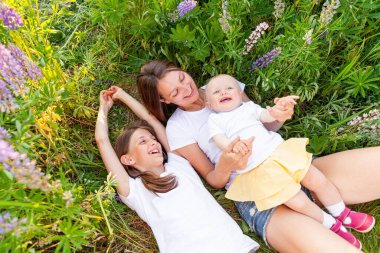 Çocuklarını kucaklayan genç bir anne. Kadın bebek ve genç kız yaz tarlasında uzanmış çiçek açan yeşil arka planda. Mutlu aile anne ve kızları çayırda oynuyorlar