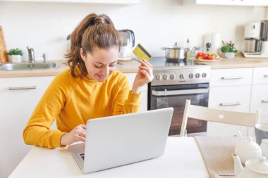 İnternetten alışveriş yapan ve kredi kartıyla ödeme yapan bir kadın. Bilgisayarla internetten alışveriş yapan genç bir kız kredi kartı bilgilerini mutfak arka planına kaydet. Çevrimiçi alışveriş e-ticaret kavramı