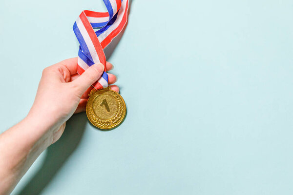 Simply flat lay design hand holding winner or champion gold trophy medal isolated on blue colorful background. Victory first place of competition. Winning or success concept. Top view copy space