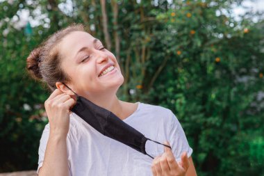 Mutlu pozitif kız koruyucu tıbbi maskeyi açık havadan çıkarıyor. Aşıdan sonra gülümseyen maskeyi çıkaran genç bir kadın. Coronavirus salgını Covid 19 konsepti. Bahar çiçekleri polen alerjisi.