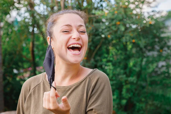 Menina positiva feliz tira máscara médica protetora do rosto ao ar livre. Mulher jovem removendo máscara sorrindo após a vacinação. pandemia de coronavírus Conceito de Covid 19. Flores da primavera pólen alergia. — Fotografia de Stock