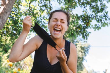 Mutlu pozitif kız koruyucu tıbbi maskeyi açık havadan çıkarıyor. Aşıdan sonra gülümseyen maskeyi çıkaran genç bir kadın. Coronavirus salgını Covid 19 konsepti. Bahar çiçekleri polen alerjisi