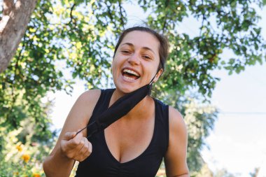 Mutlu pozitif kız koruyucu tıbbi maskeyi açık havadan çıkarıyor. Aşıdan sonra gülümseyen maskeyi çıkaran genç bir kadın. Coronavirus salgını Covid 19 konsepti. Bahar çiçekleri polen alerjisi