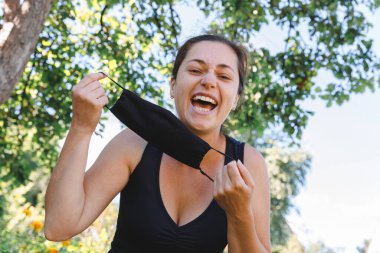 Mutlu pozitif kız koruyucu tıbbi maskeyi açık havadan çıkarıyor. Aşıdan sonra gülümseyen maskeyi çıkaran genç bir kadın. Coronavirus salgını Covid 19 konsepti. Bahar çiçekleri polen alerjisi