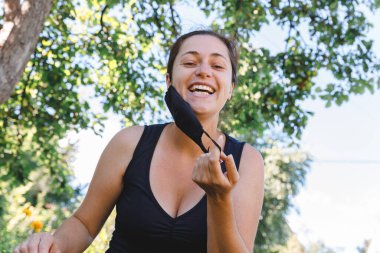 Mutlu pozitif kız koruyucu tıbbi maskeyi açık havadan çıkarıyor. Aşıdan sonra gülümseyen maskeyi çıkaran genç bir kadın. Coronavirus salgını Covid 19 konsepti. Bahar çiçekleri polen alerjisi