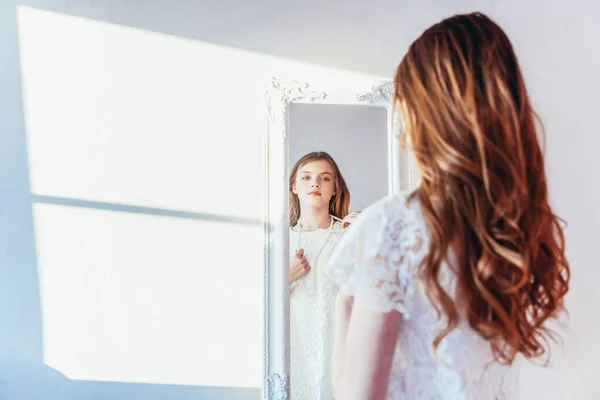 Beauty makeup morning rutine love yourself concept. Young teenage girl looking at reflection in mirror. Young positive woman wearing white dress posing in bright light room against white wall