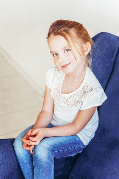 Sweet little girl in jeans and white T-shirt sitting on modern cozy blue chair relaxing in white bright living room at home indoors. Childhood schoolchildren youth relax concept