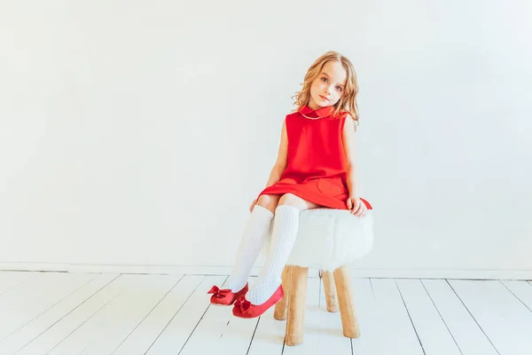 白い明るいリビングルームに屋内でリラックスし 自宅で白い壁に対して椅子に座って赤いドレスの甘い女の子 子供の頃の若者のリラックスコンセプト — ストック写真