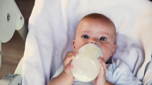 ボトルからミルクを飲んで、白い背景のカメラを見てかわいい新生児の女の子。乳児は自宅の給餌椅子に横になってミルクの栄養を食べて吸う。母親の幸せな子供 — ストック動画