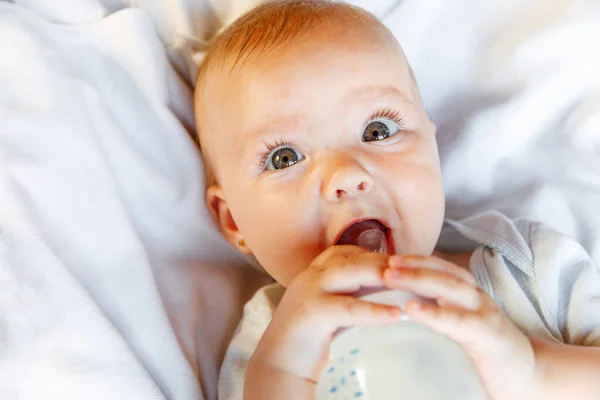 Мила маленька новонароджена дівчинка п'є молоко з пляшки і дивиться на камеру на білому тлі. Дитина, яка їсть молоко, лежить на ліжку вдома. Батьківство щаслива дитяча концепція . — стокове фото