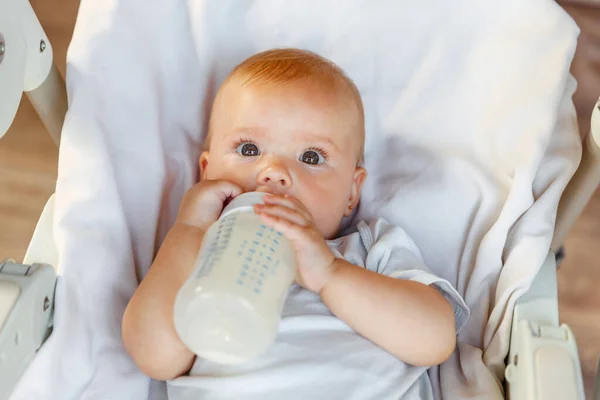 Aranyos újszülött lány tejet iszik üvegből, és fehér háttérrel nézi a kamerát. A csecsemő csecsemő szopogatja a tejet, miközben otthon fekszik az etetőszéken. Anyaság boldog gyermek — Stock Fotó