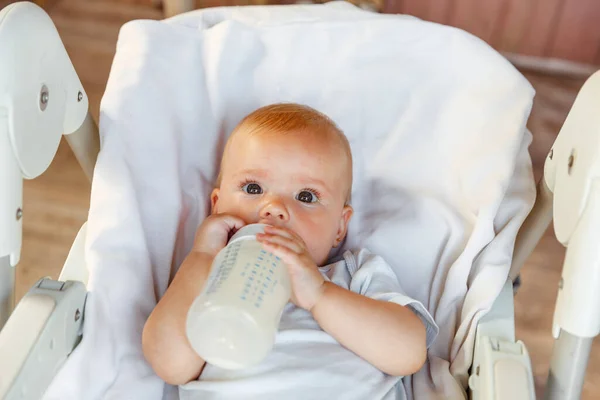 Cute little newborn girl drinking milk from bottle and looking at camera on white background. Infant baby sucking eating milk nutrition lying down on feeding chair at home. Motherhood happy child — Stock Photo, Image
