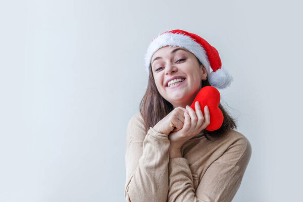 Beautiful girl in red Santa Claus hat holding red heart in hand isolated on white background. Young woman portrait, true emotions. Happy Christmas and New Year holidays concept